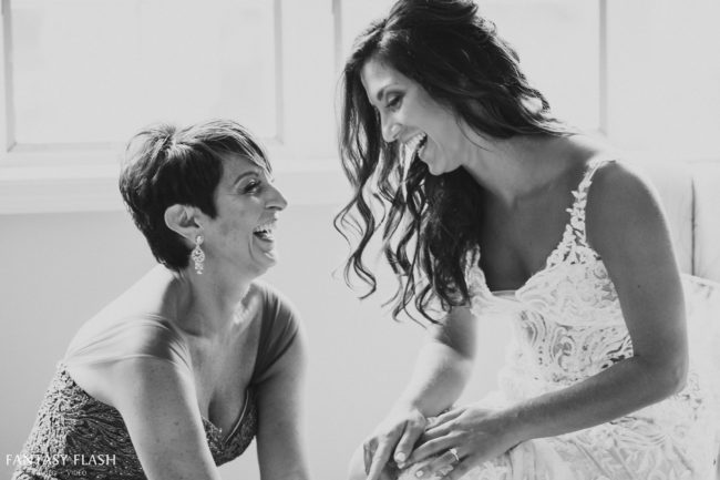 i-m3M9TPH-X4 This is a black and white photo of a mother and daughter laughing together while getting ready for the daughter's wedding day. The mother is helping the daughter put on her wedding dress.